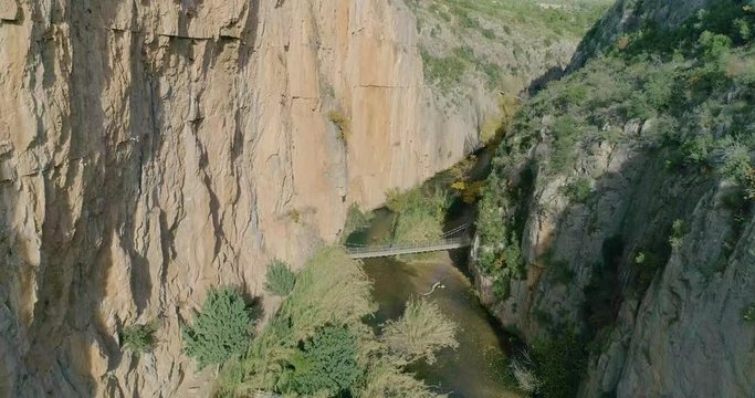 Dron se acerca lentamente dentro de barranco de paredes verticales y un puente colgante al fondo con senior cruzando