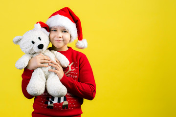 9 year old boy in a red Santa hat is holding a white soft  bear of a friend in his arms on a yellow background in the studio. New Year and Christmas concept.Copy space.