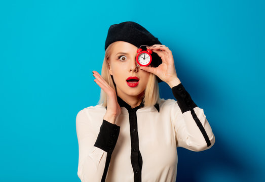 beautiful french woman in beret holds red alarm clock on blue background