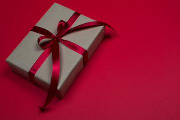 Gift box on a red background. Satin burgundy ribbon, bow, box wrapped in craft paper. Luxury gift, place for text. Holidays concept. View from above, layout for design.