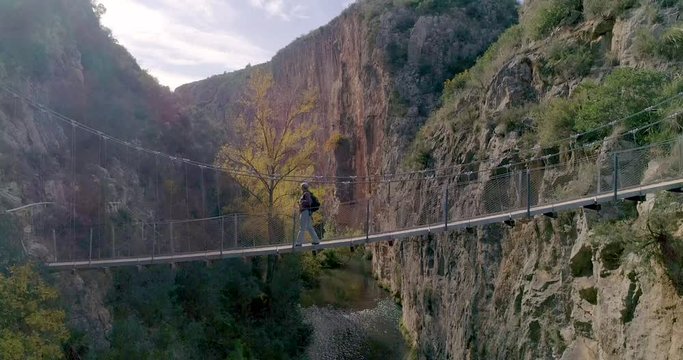 Barranco de paredes verticales y jubilado con canas cruzando puente colgante y el dron ascendiendo