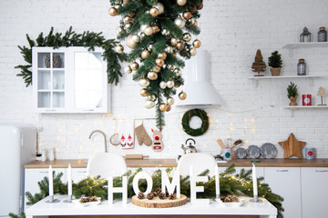 Interior of minimalistic kitchen with white walls, white floor, white countertops. Kitchen with white furniture. Kitchen decorated with  garlands and Christmas toys. Christmas tree upside down.