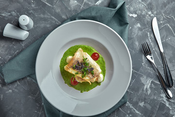 White steak of grilled fish on pillow of vegetables and yellow sauce, with tomatoes and fresh herbs. Fish on white plate with white wine on dark beautiful background. Close-up. Space