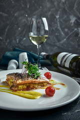 White steak of grilled fish on pillow of vegetables and yellow sauce, with tomatoes and fresh herbs. Fish on white plate with white wine on dark beautiful background. Close-up. Space