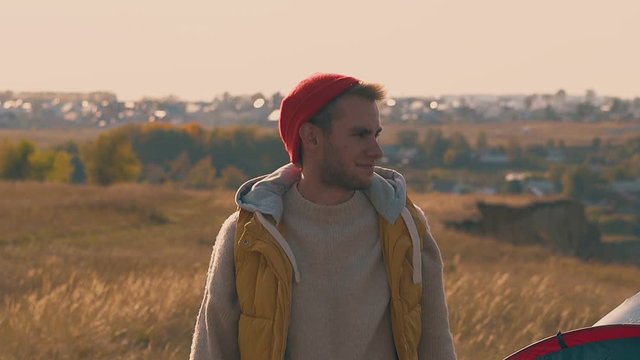 smiling guy fixes orange hat and walks away on yellow meadow against blurry village house silhouettes slow motion
