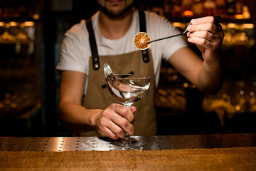 Professional male bartender adding to an alcoholic cocktail a dried lemon slice with tweezers