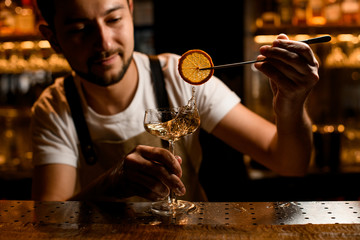 Bartender adding to an alcoholic cocktail a dried lemon slice with tweezers