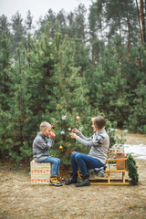 Pretty young mother and the son in gray sweaters decorating a Christmas tree in winter forest outdoors. Boy is drinking hot tea and sitting on wooden sledge with present boxes under the tree