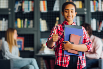 Knowledge, education and school concept, artican woman holding  book iand fplders n library, okay sign.