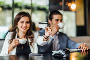 Portrait of young prety woman holding credit card at hands while man drinking coffee. Payment concept.
