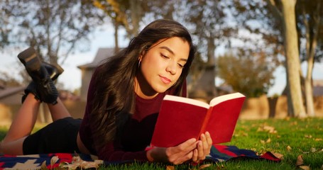 A young hispanic school girl reading a book and studying in the park before her college literature class. - Powered by Adobe