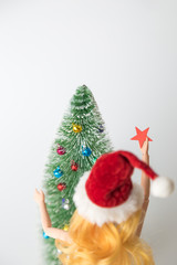 Rear view of girl doll in Santa hat holding star decoration next to fir tree and preparing for Christmas home decoration minimal creative holiday concept.
