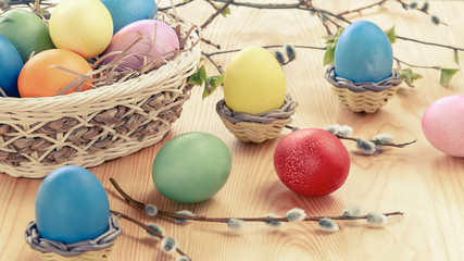 Easter composition - several colored eggs in a basket and on a light wooden table with willow twigs