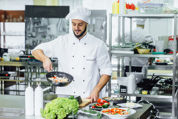 Chef with ckicken and other food in the kitchen, handsome young man with apron frying vegetables for pasta.