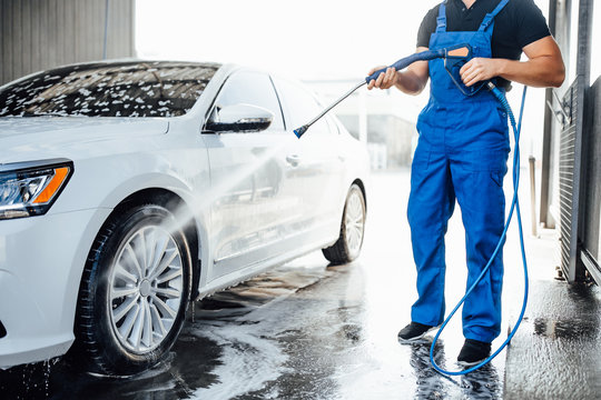 Professional washer in blue uniform washing luxury car with water gun on an open air car wash. Close up photo.