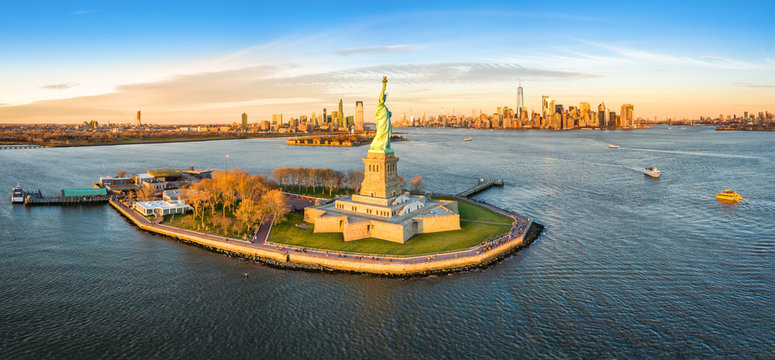 Aerial panorama of the Statue of Liberty in front of Jersey City and New York City skylines at sunset.