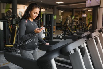 Fototapeta na wymiar Young fit woman running on treadmill listening to music via headphone at gym. Concept of healthy lifestyle.
