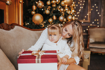 Fototapeta na wymiar Young blonde mother and her daughters in white knitted clothes opening a magical Christmas gift by a Christmas tree in cozy living room in winter