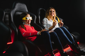 Fototapeta na wymiar Happy mother and daughter wearing 3d glasses in cinema. Cheerful family watching funny film and enjoying spare time together in movie house. Concept of enjoyment and fun.