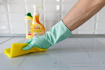 Close up of man hand in protective rubber gloves washing or cleaning modern white induction hob by a rag in the kitchen, blurred cleaning products on background. 