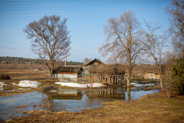 Russian village. An old rustic wooden house behind a low wooden picket fence in spring. Spring puddles on the background of a village hut in Russia.