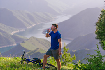 Fototapeta na wymiar Mountain biker drinks water while standing at a green valley and having some rest.