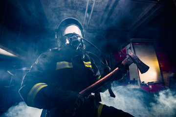Obraz na płótnie Canvas Portrait of a female firefighter while holding an axe and wearing an oxygen mask indoors surrounded by smoke.
