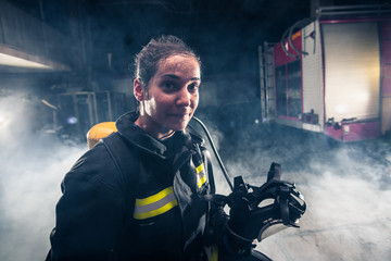 Portrait of a female firefighter while holding an axe and wearing an oxygen mask indoors surrounded...