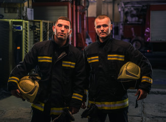 Portrait of two young firemen in uniform standing inside the fire station .