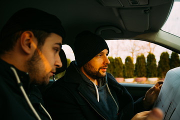 Two armed bandits sitting in a car planning their next robbery, while counting on a stopwatch the time they have for the robbery, showing off their guns and pointing at the blueprint.