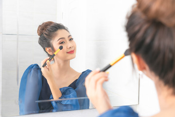 Young asian woman with makeup brush and highlighting palette looking to mirror at bathroom.