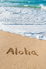 The word Aloha written in the sand on the beach with a wave washing in