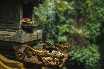 Traditional balinese Canang Sari offerings to gods and spirits with flowers, food and smoky aromatic sticks on dark green background. Indonesian culture and religion. Bali authentic travel concept. - obrazy, fototapety, plakaty