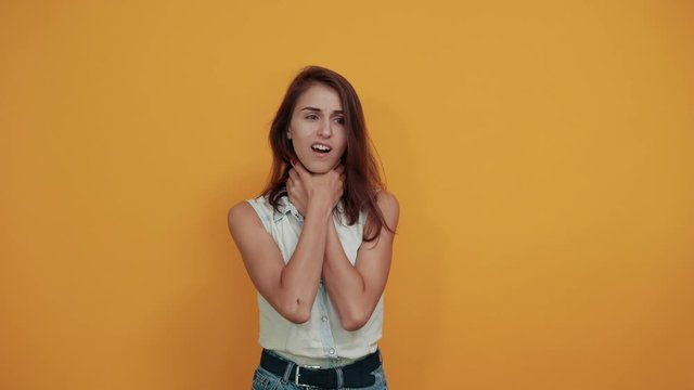 Serious caucasian young woman in blue denim shirt keeping hand on neck, strangle herself isolated on orange background in studio. People sincere emotions, lifestyle concept.