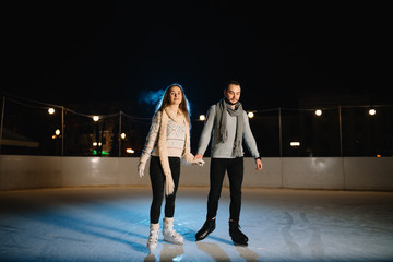 Man and woman young family Happy smiles hold hands skate on winter rink at night, with bokeh lights.