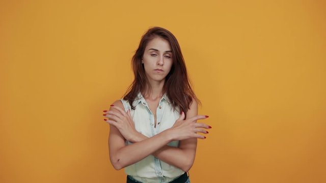 Disappointed caucasian young woman in blue denim shirt looking strict,keeping hands crossed, holding it on waist isolated on orange background in studio. People sincere emotions, lifestyle concept.