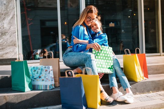 Beautiful Mom And Her Cute Little Daughter Are Holding Shopping Bags, Looking At Camera And Smiling While Standing Outdoors. Shopping Concept.