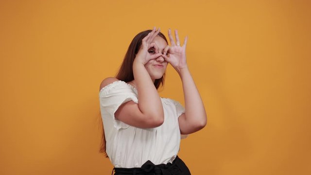 Attractive caucasian young woman in white shirt doing binoculars gesture isolated on orange background in studio. People sincere emotions, lifestyle concept.