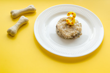 Birthday treat for puppy concept. Dog feed, candle in shape of number three, bones on yellow background copy space
