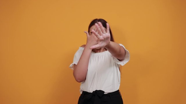 Funny young woman doing stop,NO watching gesture, holting hands on eyes, isolated on orange background in studio in casual white shirt. People sincere emotions, lifestyle concept.
