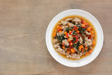 top view of stir fried pork with holy basil in a ceramic dish on wooden table. hot and spicy thai style food menu.