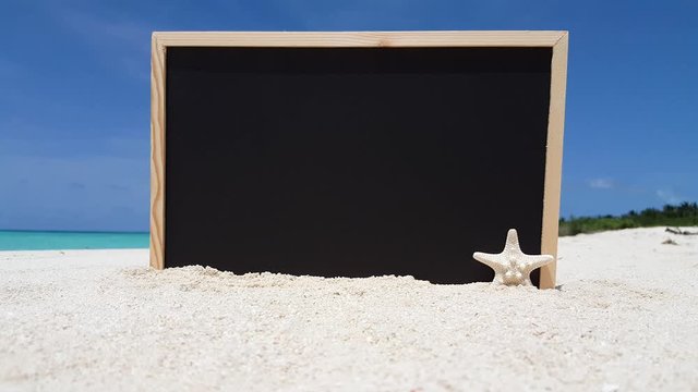Blackboard plunged on white sand of exotic beach with a blur background of sea, island and clear blue sky, copy space