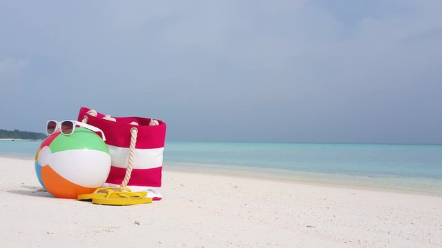 Summer bag, white sunglasses, colorful beach ball and yellow flip flops left over white sand of quiet beach on a blur background of sea and sky, copy space