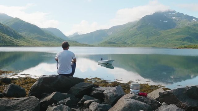 Scenic medium wide 4K static shot of male traveler hiker drinking cup of coffee from steaming camping stove, with calm lake, boat and valley of mountains in background, in Lofoten Nordland Norway.