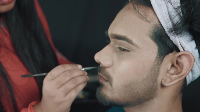 young man in cosmetics studio and professional make up artist fixing his beard with make up brush 