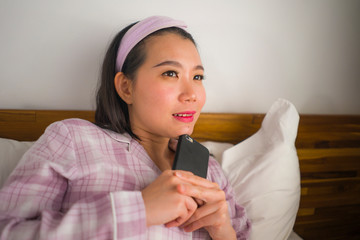 young beautiful and sweet Asian Chinese woman in cute pajamas  playful in bed using mobile phone enjoying social media or online dating app happy and relaxed