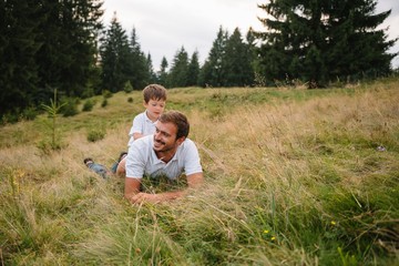 Naklejka na ściany i meble Happy father with his young son sitting on the grass on a background of green forest, mountains and sky with clouds. Friendship concept. Father with his son hiking. Father's Day