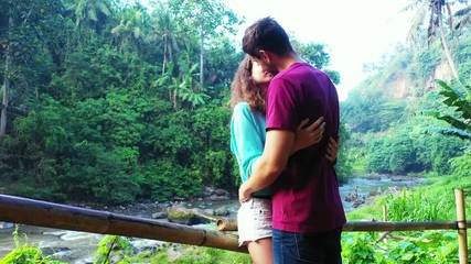 Young couple adoring and kissing each other on a beautiful tropical forest with lush vegetation and palm trees around a river valley in Thailand