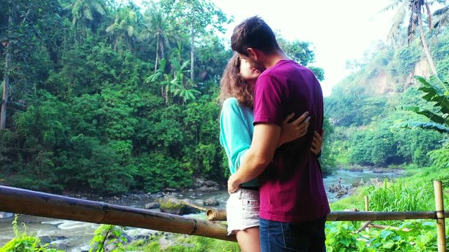 Young couple adoring and kissing each other on a beautiful tropical forest with lush vegetation and palm trees around a river valley in Thailand