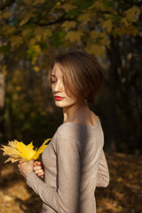 Happy girl with blonde short hair and a beautiful smile in a beige dress holds a bouquet of autumn leaves in a beautiful red-gold autumn park